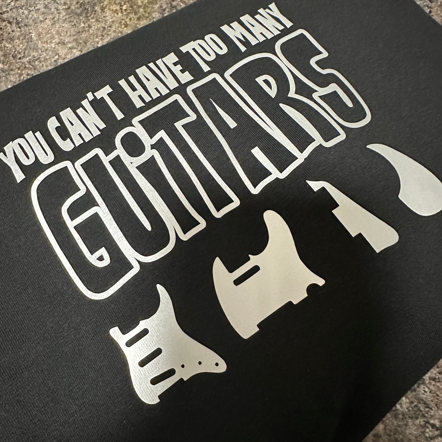 black tshirt with light grey text saying 'you can't have too many guitars' and 4 scratchplates beneath the text consisting of a Strat, tele, les Paul and acoustic and a picture of a Gibson les Paul on the right in the background at an angle, you can see some of the neck and the rhythm pickup