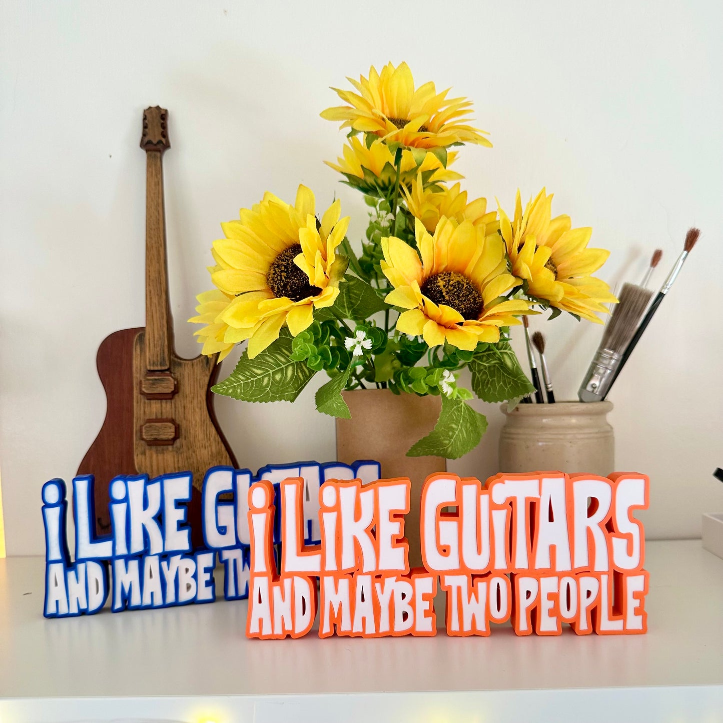 Guitar statement piece over two lines of 3d printed text one with with orange outline, the other with blue outline and white text that says 'I LIKE GUITARS' on the top line and the bottom line says 'AND MAYBE TWO PEOPLE' standing on a white shelf with a green pen pot behind and a bunch of small sunflowers to the far left in a brown cardboard tube