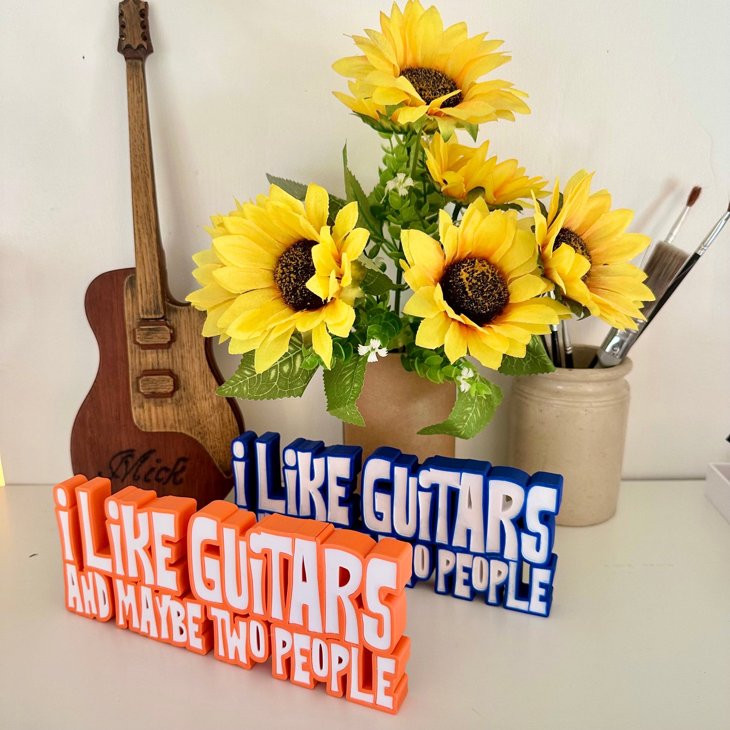Guitar statement piece over two lines of 3d printed text one with with orange outline, the other with blue outline and white text that says 'I LIKE GUITARS' on the top line and the bottom line says 'AND MAYBE TWO PEOPLE' standing on a white shelf with a green pen pot behind and a bunch of small sunflowers to the far left in a brown cardboard tube