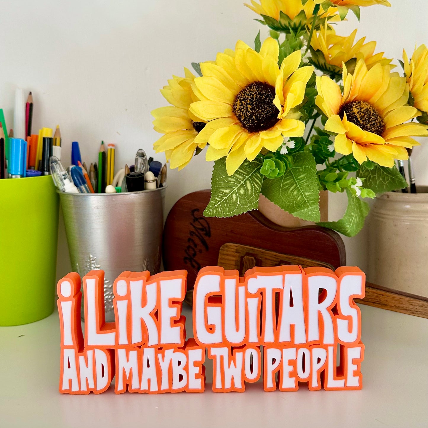 Guitar statement piece over two lines of 3d printed text with with orange outline, and white text that says 'I LIKE GUITARS' on the top line and the bottom line says 'AND MAYBE TWO PEOPLE' looking at it straight on, standing on a white shelf with a green pen pot behind and a bunch of small sunflowers to the far right in a brown cardboard tube