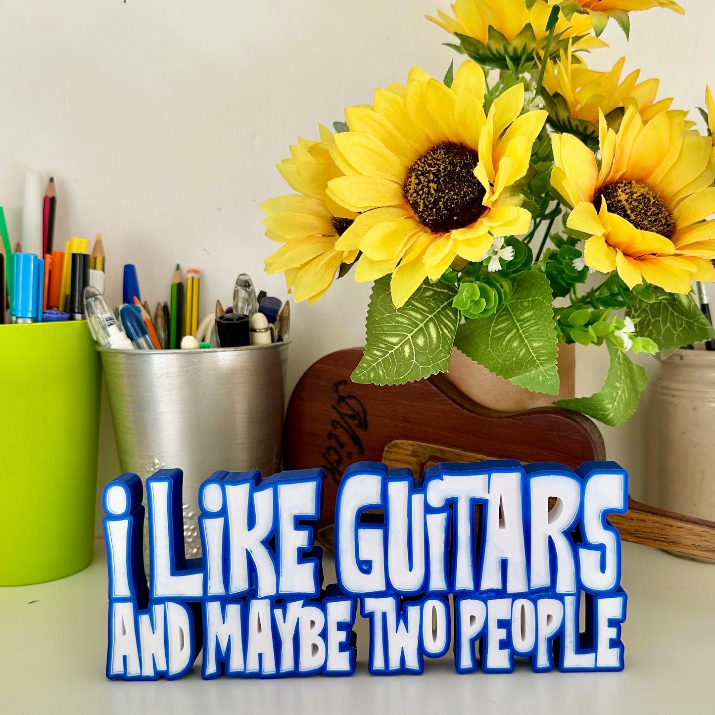 Guitar statement piece over two lines of 3d printed text with with a blue outline, and white text that says 'I LIKE GUITARS' on the top line and the bottom line says 'AND MAYBE TWO PEOPLE' looking at it from the left angle, standing on a white shelf with a green pen pot behind and a bunch of small sunflowers to the far left in a brown cardboard tube