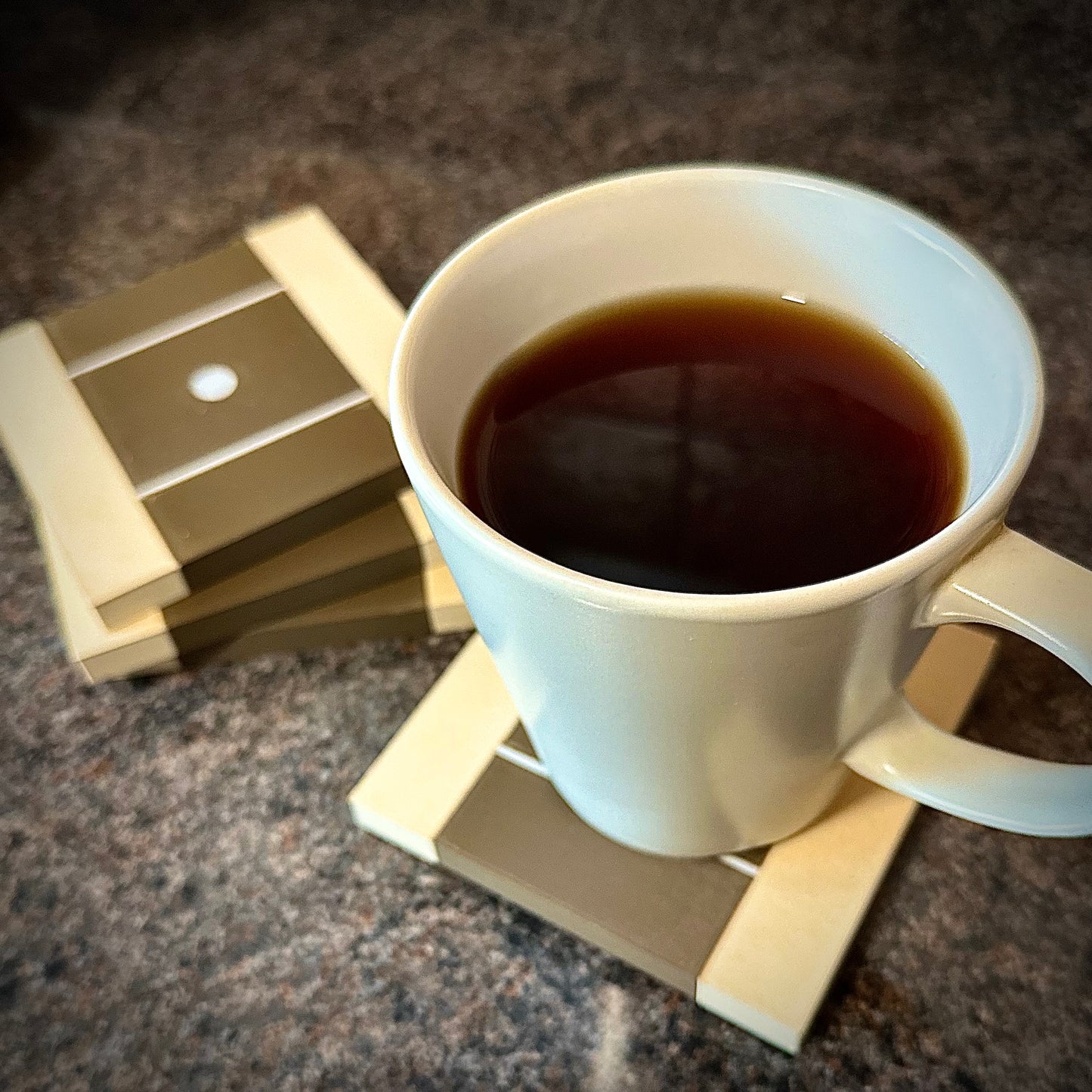4 square guitar neck Coasters 4" 100mm square in size with light maple colour edges and walnut colour in the middle to signify the guitar fretboard with Stratocaster dot inlay. Foreground demonstrated cup on the coster with the other 3 to the side with a slight bokeh