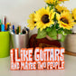 Guitar statement piece over two lines of 3d printed text with with orange outline, and white text that says 'I LIKE GUITARS' on the top line and the bottom line says 'AND MAYBE TWO PEOPLE' looking at it straight on, standing on a white shelf with a green pen pot behind and a bunch of small sunflowers to the far right in a brown cardboard tube