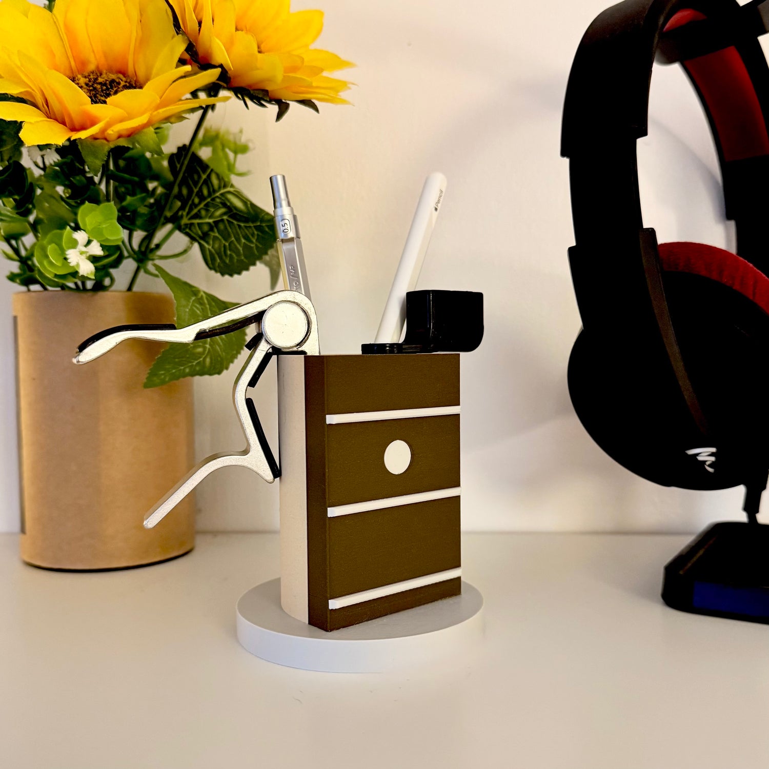 Dot inlay guitar neck pen holder standing on white shelf containing a cap, string winder, pen and Apple Pencil. To the right side are Focal headphones and to the left a brown plant pot with sunflowers in. Background is white